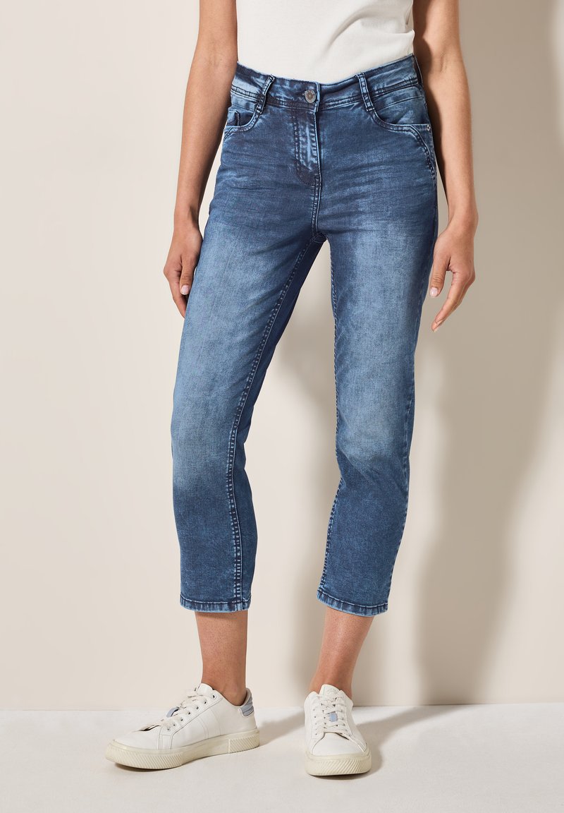Jean en denim avec une finition bleue délavée, taille mi-haute, longueur cropped et design classique à cinq poches, associés à des baskets blanches.