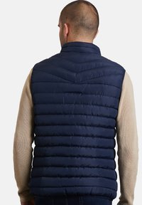 Marineblauer Puffer-Weste mit gestepptem Design, vertikalen Streifen und Stehkragen, die über einem beigen Strickpullover getragen wird.