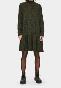 Mörkgrön klänning med ett mönstrat tröjmaterial, hög krage, långa ärmar och flerskiktad nederdel, tillsammans med svarta snörade ankelstövlar.