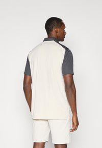 Camisa polo en beige con mangas cortas en gris oscuro, con dobladillo recto y cuello. El material parece suave y ligero.