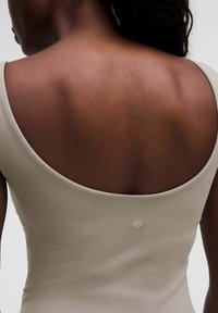 Beige mouwloze jumpsuit met een laag rugontwerp, voorzien van een gladde textuur en een subtiele logo in het midden nabij de schouder.
