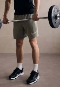 Shorts sportivi verdi con un logo Nike stampato, abbinati a sneakers nere e calze bianche. Bilanciere tenuto in mano, gambe muscolose visibili.