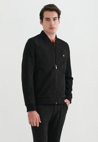 Sort ruskinds bomberjakke med lynlås fortil, ribbede manchetter og et lille logo på brystet. Bæres over en orange skjorte, kombineret med mørke bukser.