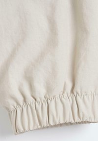 Tissu beige clair avec un ourlet élastiqué froncé, montrant des détails de texture et de couture.