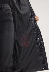 Czarna ocieplana kurtka puchowa o błyszczącej, gładkiej fakturze. Wewnątrz znajduje się nadrukowana etykieta pielęgnacyjna. Z przodu zapinana na zatrzaski.