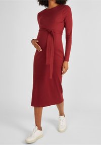 Schwangerschaftskleid aus rotem Rippstoff, mit langen Ärmeln und einem Bindegürtel in der Taille. Getragen mit weißen Sneakern. Mittellanges Design.