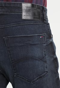 Ciemnoniebieskie dżinsy o teksturowanej powierzchni, z skórzaną naszywką z napisem "TOMMY JEANS" oraz małą flagą na tylnej kieszeni.