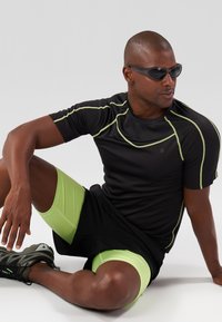 T-shirt sportivo nero con accenti verdi, abbinato a pantaloni corti verdi e scarpe da ginnastica grigie. La figura indossa occhiali da sole scuri.