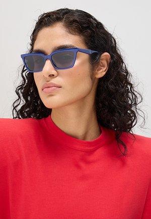 Donna con capelli scuri e ricci che indossa occhiali da sole rettangolari blu e una maglietta rossa vivace, rivolta leggermente a sinistra su uno sfondo chiaro e semplice.