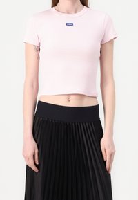 Kvinde iført en lys rosa cropped t-shirt med "HUGO" logo og en højtsiddende sort plisseret nederdel, stående foran en hvid baggrund.