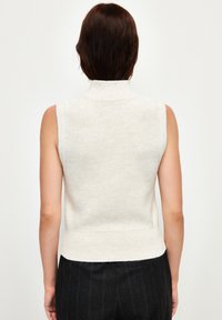 Top a colloque alto senza maniche in beige chiaro. Tessuto morbido con orlo a coste, presenta una vestibilità comoda. Indossato su pantaloni scuri a righe.