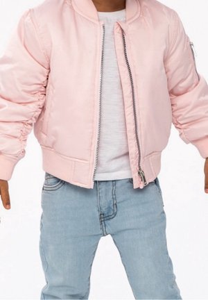 Barn iført lys pink bomberjakke over hvid skjorte og lyseblå jeans, stående med lidt udstrakte arme.