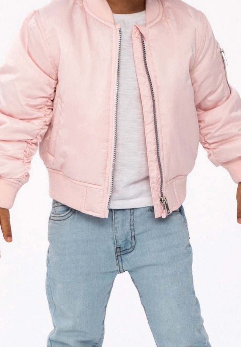 Enfant portant une veste bomber rose clair sur une chemise blanche et un jean bleu clair, debout avec les bras légèrement écartés.