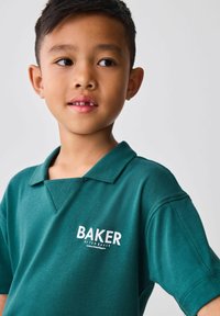 Mladý chlapec v zelenom košelovom tričku s nápisom "BAKER by Ted Baker", ktorý sa mierne pozerá na stranu pred jednoduchým pozadím.