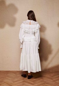 Λευκό φόρεμα με δαντελένιο γιακά, μακριά φουσκωτά μανίκια και πολυεπίπεδη φούστα. Φορεμένο με σκούρα παπούτσια πάνω σε ξύλινο πάτωμα μπροστά σε υφασμένο φόντο.