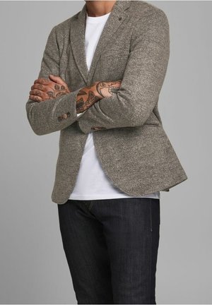 Homme portant un blazer chevrons sur un t-shirt blanc, bras tatoués croisés et jeans foncés sur fond gris.