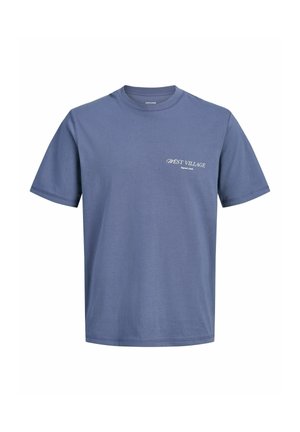 Dreng smilende med kort mørkt hår, iført en blå T-shirt, lyseblå jeans og beige sko, stående mod en ensfarvet hvid baggrund.