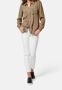 Camicetta marrone chiaro a maniche lunghe con dettaglio arricciato sul davanti, abbinata a jeans skinny bianchi e sneakers nere. L'outfit casual mette in risalto un design minimalista.