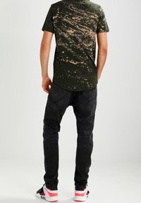 Camiseta verde con un patrón moteado beige, combinada con vaqueros negros y zapatillas con detalles en rojo. Vista trasera del conjunto.