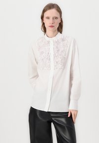 Chemisier blanc à boutons avec un motif floral texturé sur le panneau avant, doté d'un col Mao et de manches longues, associé à un pantalon noir.