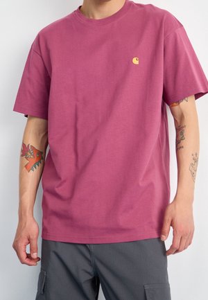 Homme portant un t-shirt uni à manches courtes rose foncé avec un petit logo jaune sur la poitrine et un pantalon cargo gris foncé.