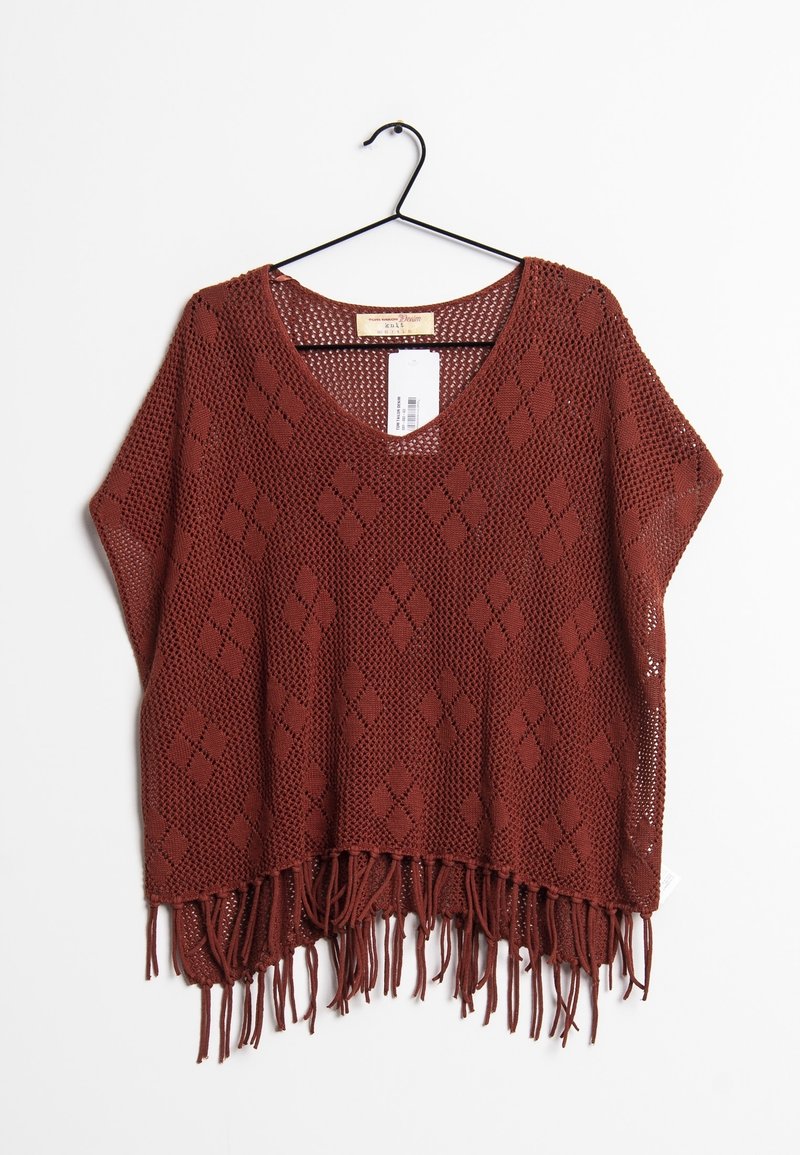 Poncho aperto color ruggine con motivo a diamante e orlo frangiato, appeso a un gancio nero su uno sfondo bianco.