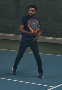 Τένις παίκτης σε ναυτικό πουκάμισο και μαύρο αθλητικό παντελόνι, κρατώντας μια πορτοκαλί ρακέτα τένις. Εσωτερικό γήπεδο με πράσινο φόντο.