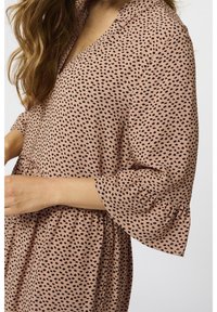 Femme portant une robe beige avec un motif de points noirs irréguliers et des manches bouffantes aux coudes, sur un fond uni.