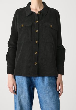 Sort kordfløjls skjorte med knapper og brystlommer båret med blå denimjeans, vist fra hals til hofter.