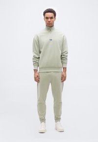 Άνδρας στέκεται φορώντας ανοιχτοπράσινο φούτερ με φερμουάρ και ταιριαστό παντελόνι φόρμας με λευκά αθλητικά, πάνω σε απλό λευκό φόντο.