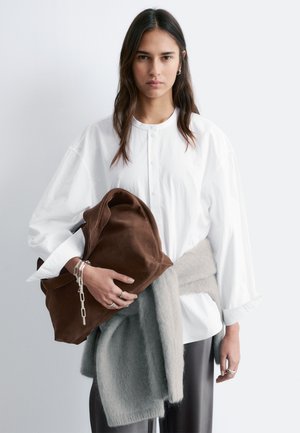 Kvinde med langt mørkt hår iført en løs hvid skjorte med knapper og en grå sweater bundet om taljen, der holder en stor brun ruskindstaske.