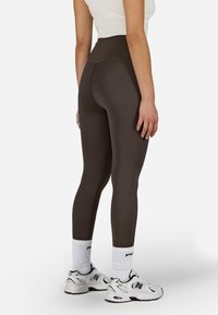 Magas derekú, sima anyagból készült barna leggings, díszvarrásokkal és feszes illeszkedéssel. Fehér bokazoknival és sportcipővel viselve.