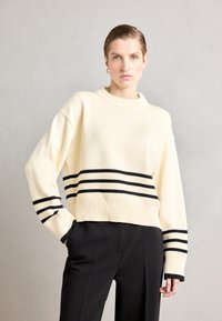 Creme trui van gebreide stof, met zwarte horizontale strepen en een geribde zoom en manchetten. G gedragen met zwarte broek.