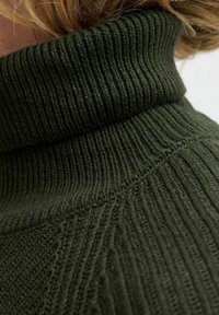 Dunkelgrüner, gestrickter Rollkragenpullover mit geripptem Design, hoher Kragen und diagonalen Nahtdetails. Nahaufnahme.