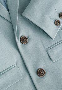 Blazer azzurro chiaro con texture sottile, bottoni marroni, revers a punta e dettagli sulle tasche. Tessuto morbido adatto per abbigliamento formale.