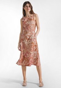 Vrouw met een mouwloze jurk tot op de knie met oranje en roze tropische bladerenpatroon, staand met één hand in de zak, beige ballerina's.