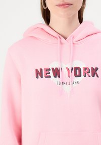Felpa rosa con tasca a marsupio, caratterizzata da una grafica "New York" in rosso e nero con contorni a forma di cuore e dettagli bianchi. Materiale in cotone misto.