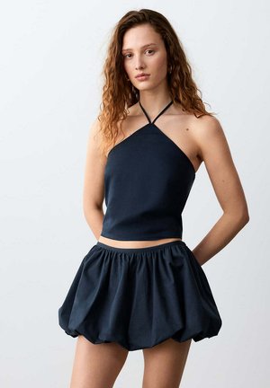 Jeune femme aux cheveux bouclés portant un haut halter noir associé à une jupe boule noire volumineuse, debout les mains derrière le dos.