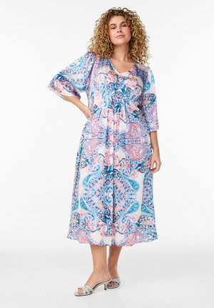 Kobieta o kręconych włosach, ubrana w kolorową midi sukienkę w paisley z rękawami 3/4 i jasnoniebieskie sandały na niskim obcasie, stojąca z ręką na biodrze.