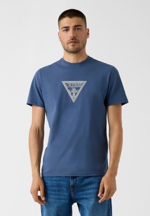 Homme portant un t-shirt bleu Guess avec un triangle de logo blanc et un jean bleu, debout devant un fond clair uni.