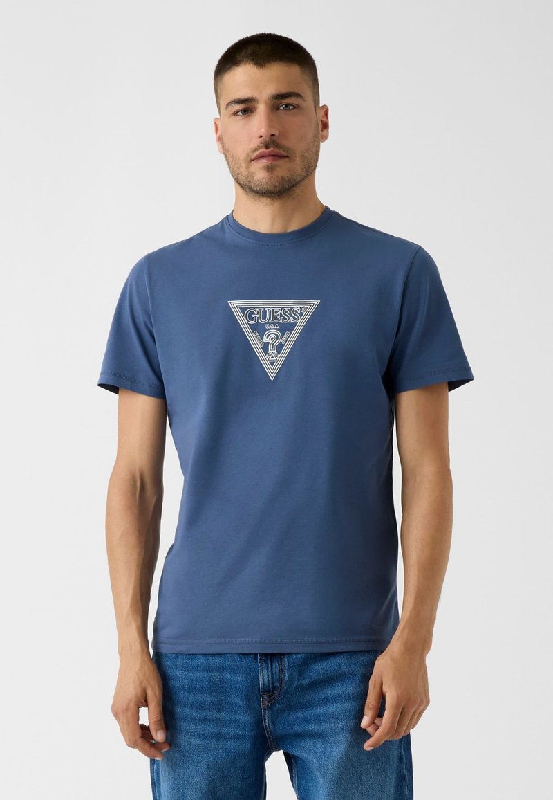 Άνδρας φορά μπλε μπλουζάκι Guess με λευκό τριγωνικό λογότυπο και μπλε τζιν, στέκεται μπροστά σε απλό φωτεινό φόντο.