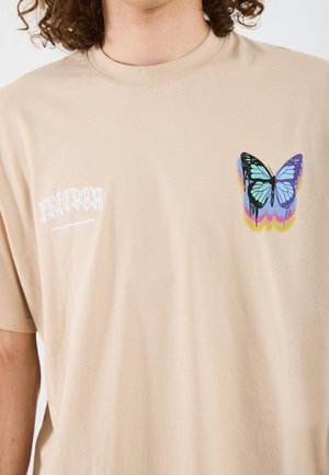 Beiges T-Shirt mit weißem "FREEDOM"-Text und einer bunten blauen Schmetterlingsgrafik im oberen Brustbereich.