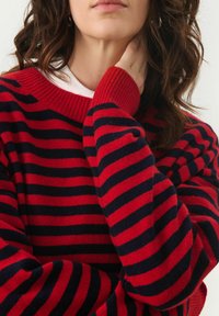 Pull en tricot rayé rouge et marine avec un col rond et des poignets côtelés, doté d'une texture douce et d'une coupe décontractée.