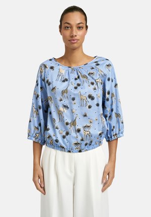 Woman wearing a light blue blouse with 3/4 sleeves featuring giraffe and palm tree patterns, paired with white pants.