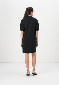 Black short-sleeve dress with a collar, featuring a relaxed fit and smooth fabric. Model is wearing black strappy high-heeled sandals.