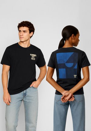 Man en vrouw dragen zwarte T-shirts en lichtblauwe spijkerbroeken, de man kijkt vooruit met tekst op de borst, de vrouw toont een geometrisch blauw ontwerp op de rug.