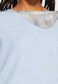 Jasnoniebieski sweter dzianinowy o miękkiej fakturze i szerokim dekolcie, nałożony na wzorzystą, jasną bluzkę z motywami kwiatowymi.