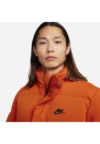 Orangen gepolsterte Jacke mit steichem Kragen, Reißverschluss vorne und Druckknopfverschluss. Verfügt über ein schwarzes Nike-Logo auf der Brust. Weicher, strukturierter Stoff.