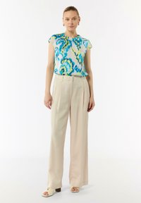 Kurzärmlige, mehrfarbige Bluse mit blauen und grünen abstrakten Mustern, kombiniert mit beigefarbenen weit geschnittenen Hosen und neutralen Riemchensandalen.