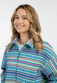 Camicia a maniche lunghe colorata, con una base turchese, strisce rosa e blu, e motivi astratti. Il modello ha capelli ondulati e una collana multicolore.